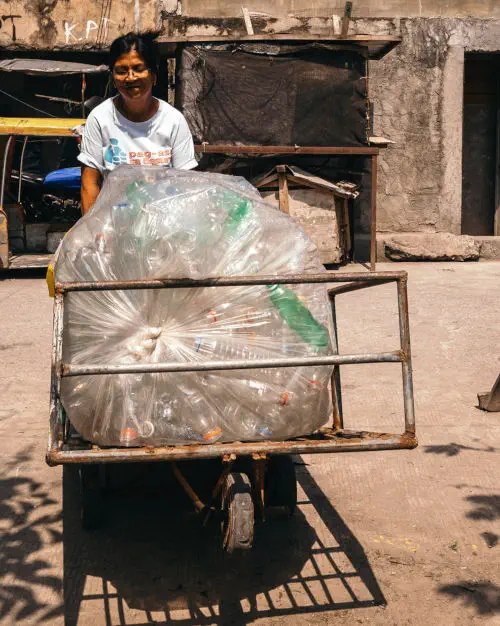 woman pushing large wheel barrow with a large bag full of plastic bottles in it